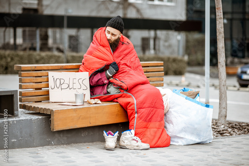 Homeless and jobless beggar sitting on the bench wrapped with sleeping bag begging money near the business center photo