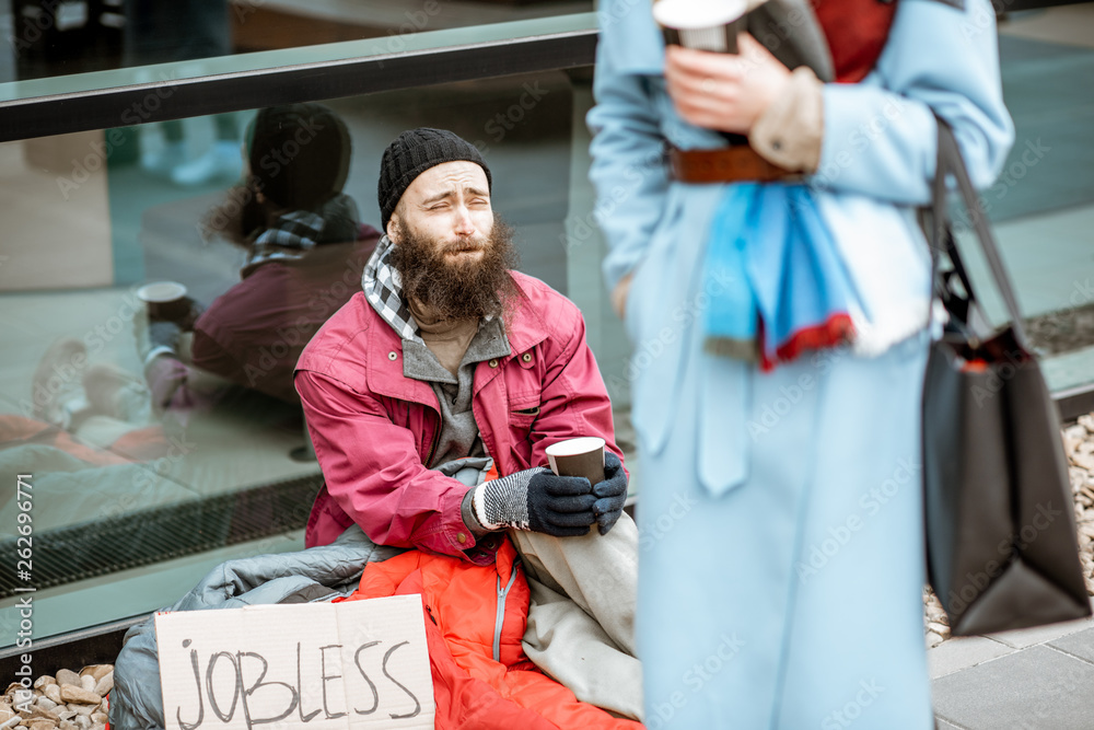 Depressed homeless begging money while sitting on the sidewalk with passing by businesswoman near the business center