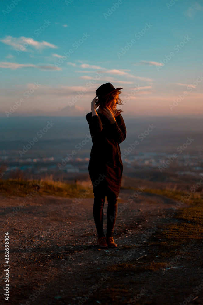girl waving her hat with her back facing the valley with mountains. aunset