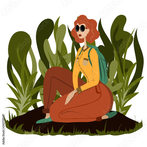 Young woman traveler with a backpack with glasses sits on the ground in the bushes.