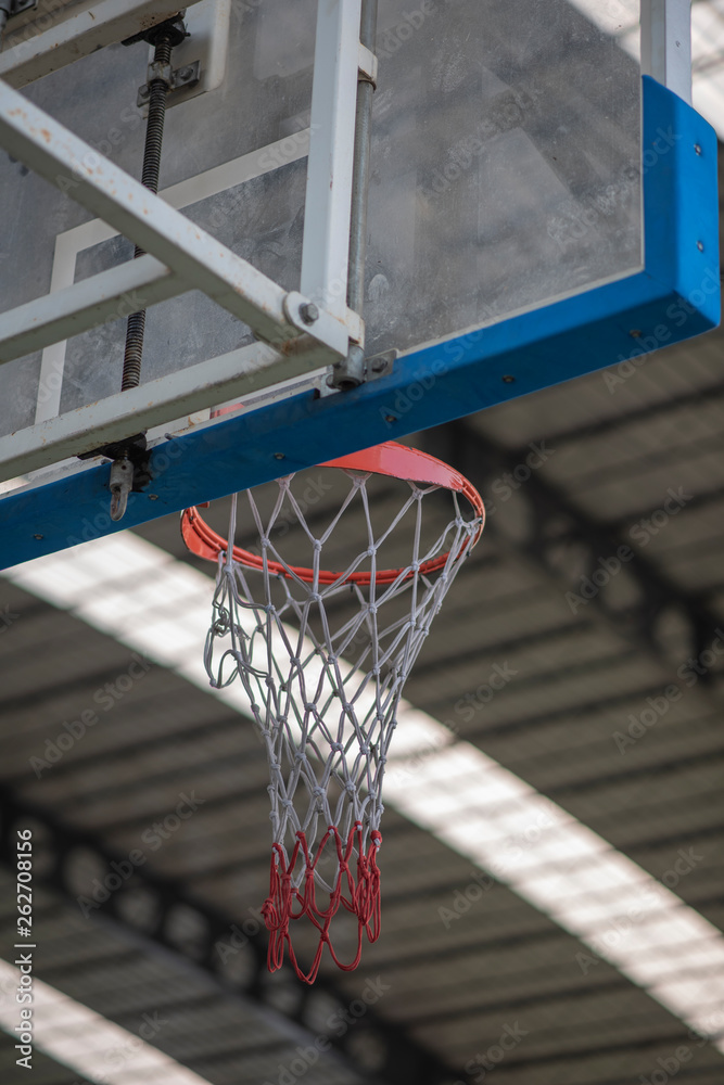 An interior space of a basket ball court showing a scaffolding metal roof sheet with a framework of metal truss with sunlight goes through and a basketball ring as a focal point.