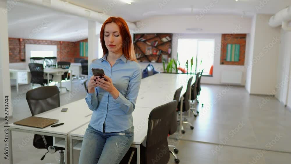 Strong independent woman in co-working space. Female worker sits on the desk while texting and smiling. Strong independent woman in co-working space. Female worker sits on the desk while texting and smiling.