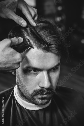 Young handsome barber making haircut for attractive bearded man at barbershop. Black and white or colorless photo. Hairstyle, salon, hairdresser, barber shop, lifestyle concept. Caucasian male models.