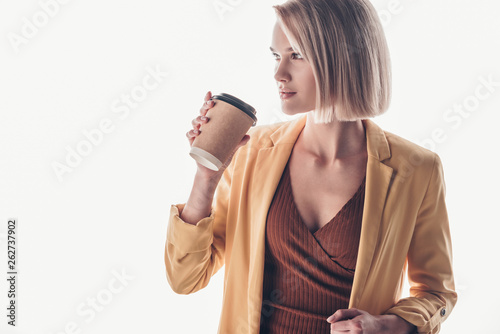 beautiful blonde woman holding disposable cup and looking away on grey