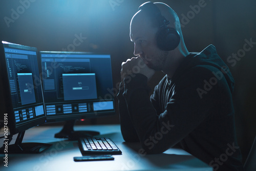 cybercrime, hacking and technology concept - male hacker in headphones with progress loading bar on computer's screens wiretapping or using computer virus program for cyber attack in dark room photo