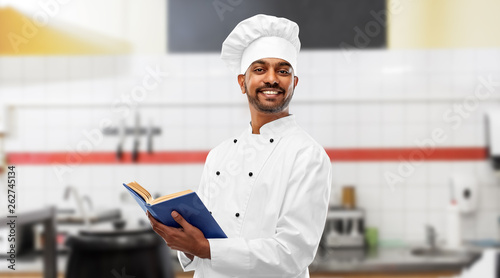 cooking, profession and people concept - happy male indian chef in toque with cookbook over restaurant kitchen background
