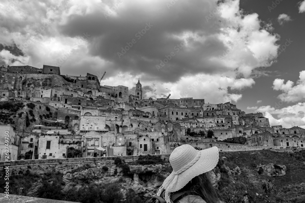 Summer day scenery view of the ancient town of the Sassi with white puffy clouds moving on Italian blue sky. Matera, Basilicata, Italy. Young woman tourist with white hat enjoys view, black and white