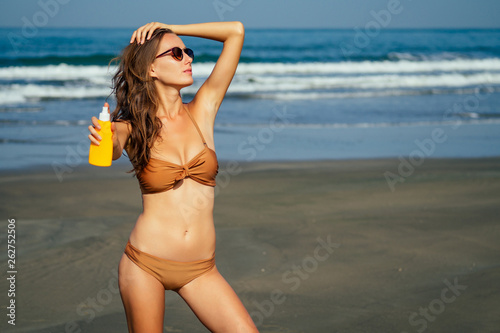beautiful tanned female fitness big boobs model holding spf bottle applying on the chest in a fashion stylish swimsuit on sea ocean beach blue sky and palm trees background.summer vacation