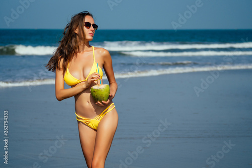 beautiful tanned girl in trendy yellow bathing suit sunglasses stands with coconut in her hands on beach tropical island vacation travel swimwear new season.derox diet idea