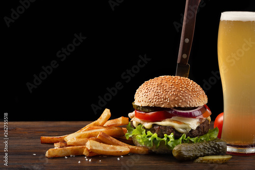 Homemade hamburger with french fries and glass of beer on wooden table. In the burger stuck a knife. Fastfood on dark background photo
