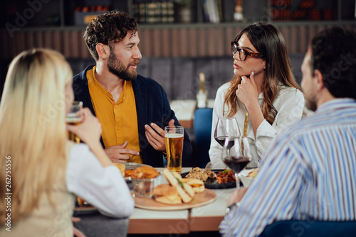 Multiethnic friends sitting at restaurant, drinking alcohol, chatting and having burgers for dinner.