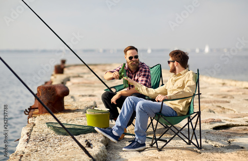 leisure and people concept - happy friends fishing and drinking beer on pier in summer