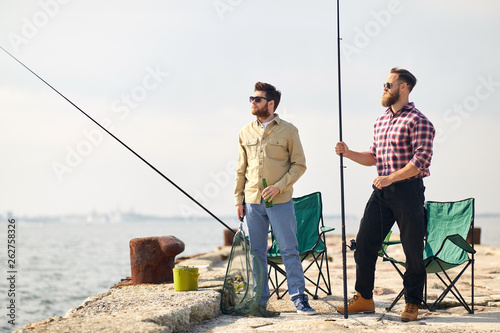 leisure and people concept - happy friends with fishing rods and beer on pier at sea