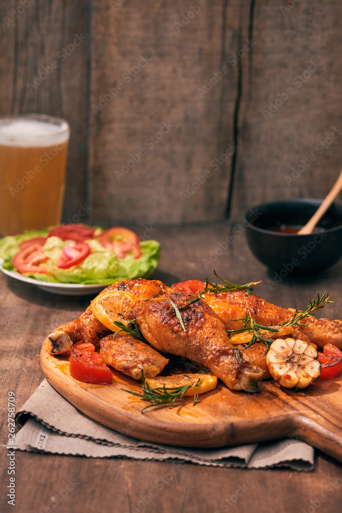 Grilled chicken legs roasted on the grill on wooden chopping board with tomato sauce in a bowl, fresh tomatoes and lettuce leaves, bitter pepper, glass mug of beer