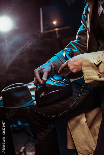 Partial view of man in trench coat using telephone in dark office
