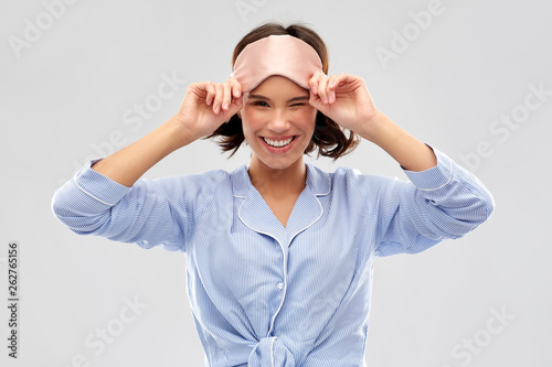 people and bedtime concept - happy young woman in pajama and eye sleeping mask winking over grey background