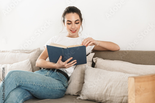 Beautiful happy young student girl sitting indoors reading book on sofa.