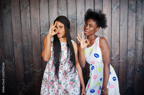 Two black african girlfriends at summer dresses posed against dark wooden background and showing okay fingers sign.
