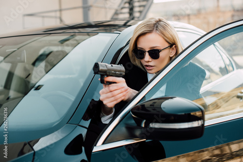 confident blonde woman in sunglasses holding gun near black car