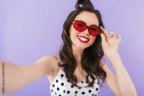 Portrait of joyful pin-up woman 20s in retro sunglasses smiling while taking selfie photo