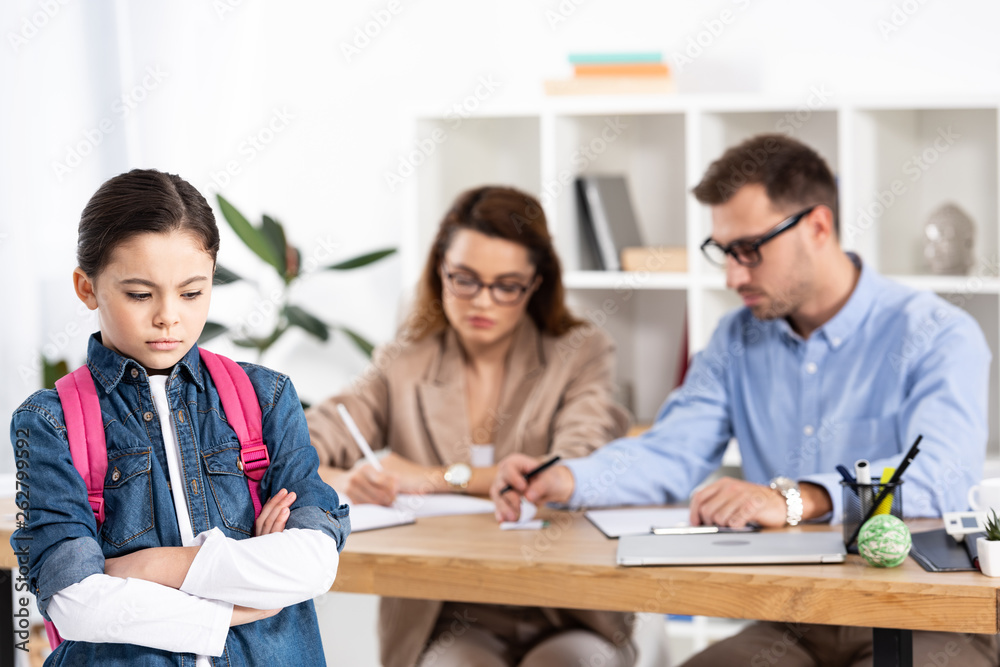 selective focus of upset kid with backpack standing with crossed arms near parents in office selective focus of upset kid with backpack standing with crossed arms near parents in office