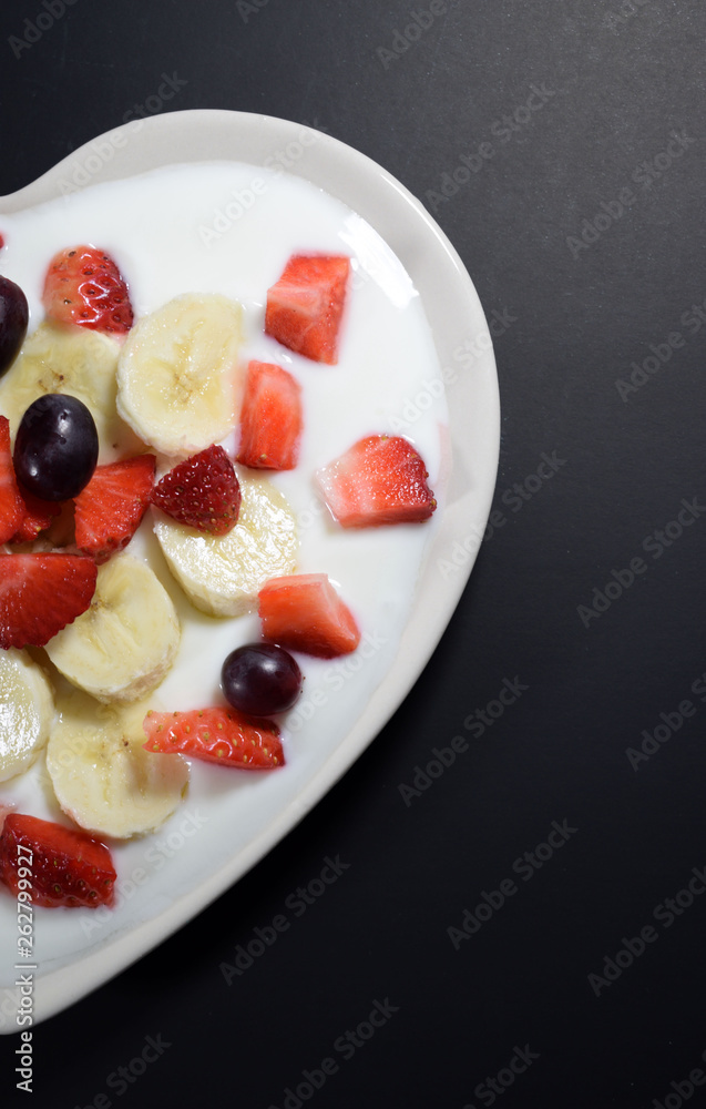 Half Heart-shaped cream plate filled with yogurt with banana slices, strawberries cut into pieces and grape berries on a black background. Half Heart-shaped cream plate filled with yogurt with banana slices, strawberries cut into pieces and grape berries on a black background.
