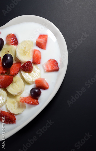 Half Heart-shaped cream plate filled with yogurt with banana slices  strawberries cut into pieces and grape berries on a black background.  Indulgence for breakfast  concept.