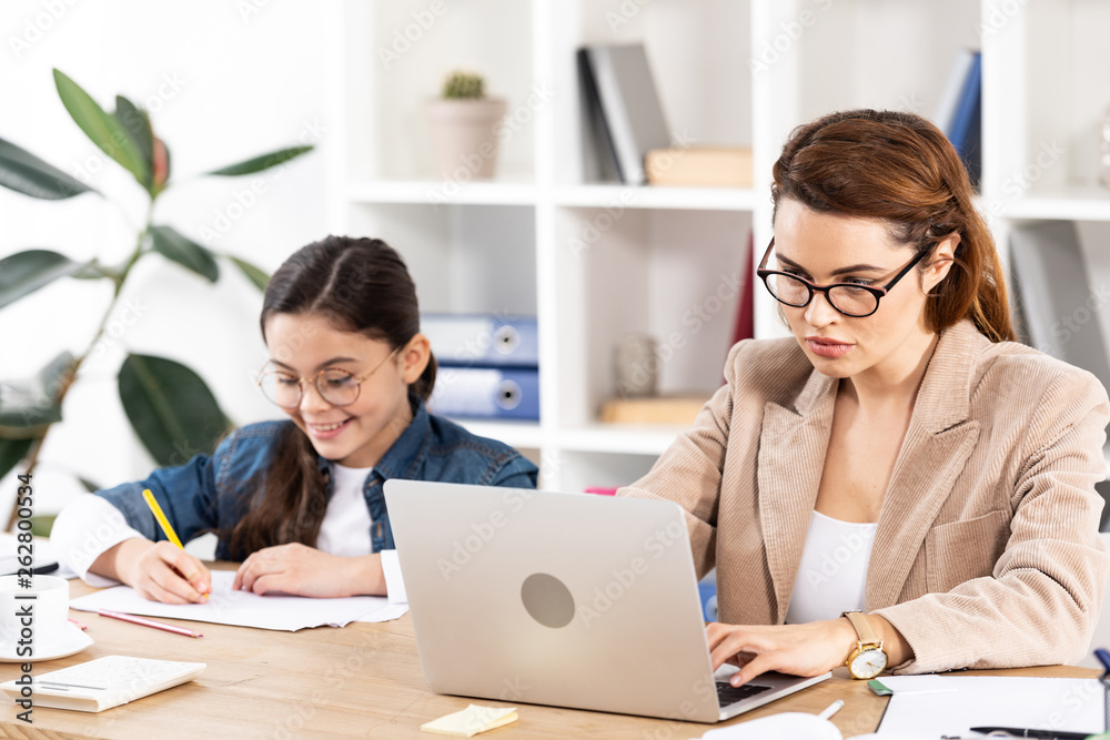 attractive mother in glasses using laptop near cheerful daughter attractive mother in glasses using laptop near cheerful daughter