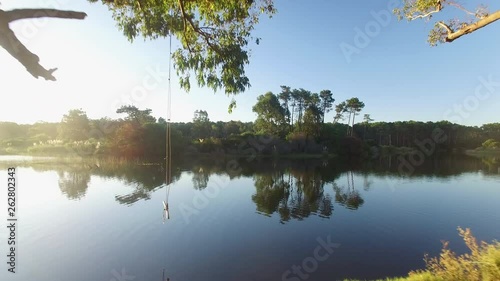 Aerial shot: flying through trees onto relective lake past a swing at sunset photo