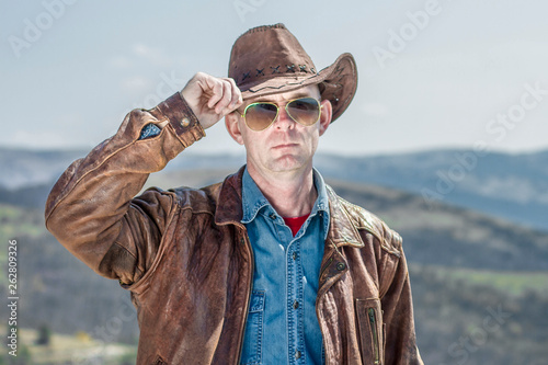 An American cowboy with glasses, a hat and a leather jacket is standing and holding his hat on his hand. Cowboy Symbol. photo