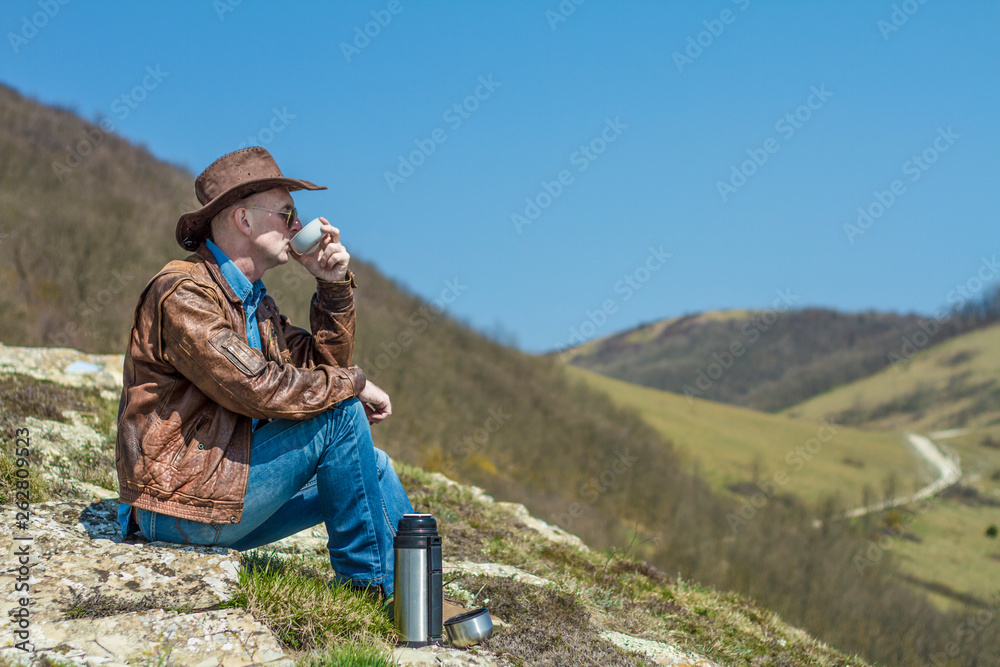 A tourist in a jacket, hat, glasses sits and drinks tea from a thermos, and looks into the distance. In the background mountains. Travel concept.