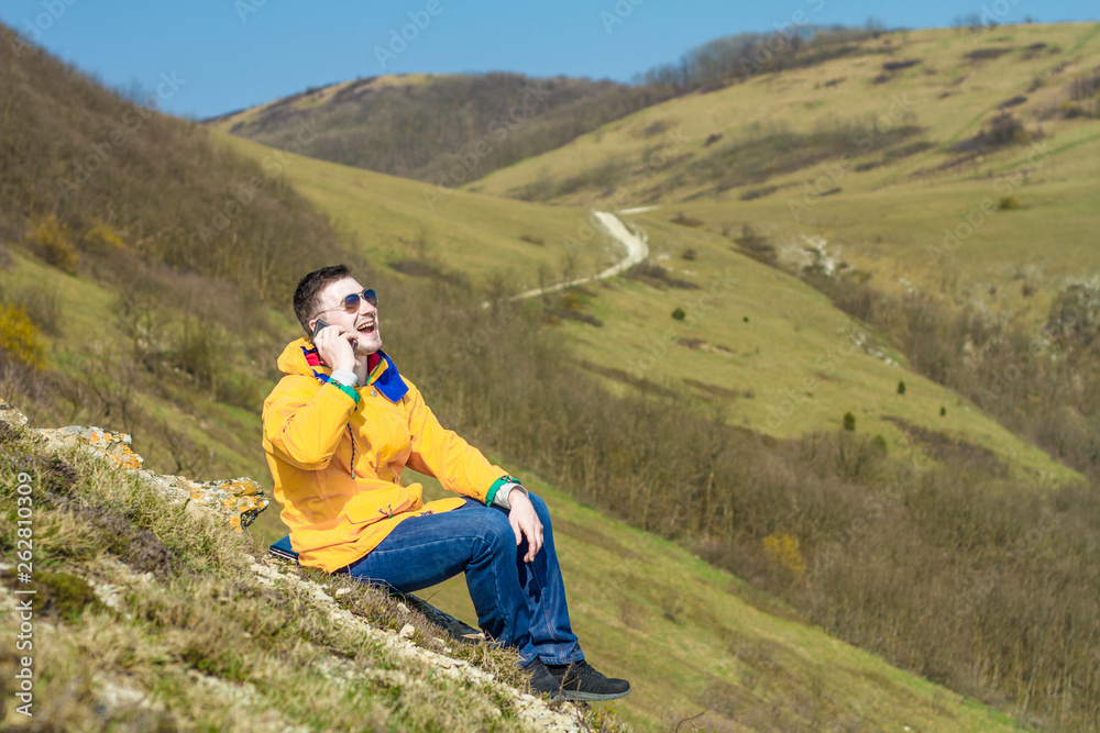 A young man in a yellow jacket, blue jeans and glasses sits in the mountains and enjoys the scenery and talking on the phone, laughing. In the background are hills and sky. Mountain landscape.
