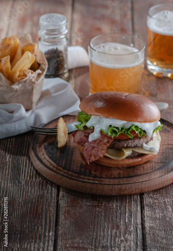  burger with bacon and cutlet with cheese, tomato, greens. served with fries and beer on a wooden table