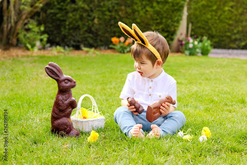 Canvas Print Cute little baby boy with bunny ears eating chocolate Easter bunnies sitting on green grass outside in the spring garden with basket of easter eggs looking at big chocolate rabbit