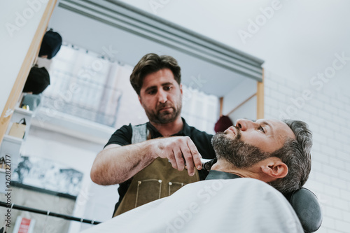 barber with comb and trimmer cutting beard of male sitting in barbershop on blurred background photo