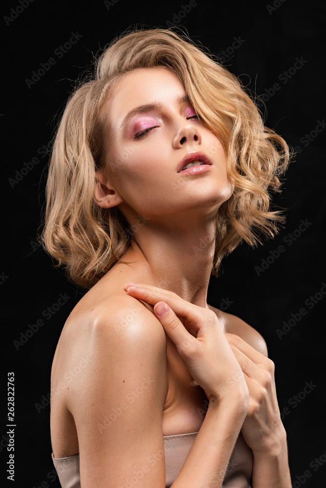 Beautiful young blonde girl with naked shoulders and curly hair, posing, with her hands sensually pressed to her breast, on a black background. Clean, healthy skin. Close-up. Beautiful young blonde girl with naked shoulders and curly hair, posing, with her hands sensually pressed to her breast, on a black background. Clean, healthy skin. Close-up.