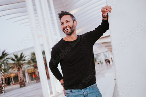 Adult handsome elegant cheerful male in casual wear leaning on grey wall and looking away on city street