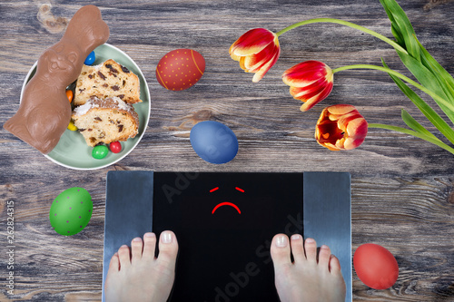 Female feet on digital scales with sad smile surrounded by Easter food (Easter cake, chocolate Easter bunny, painted eggs) and spring flowers. Concept of overeating and during holidays.