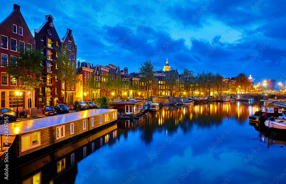 Evening town Amsterdam in Netherlands on bank river canal Amstel with shining window. Panorama landscape brown house over water reflection.