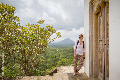 Female traveler with backpack looking away while leaning on wall of ancient oriental temple against cloudy sky in Sri Lanka