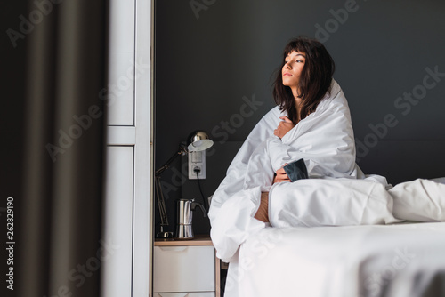 Side view of young dozy woman with mug of drink in duvet sitting on bed in bedroom