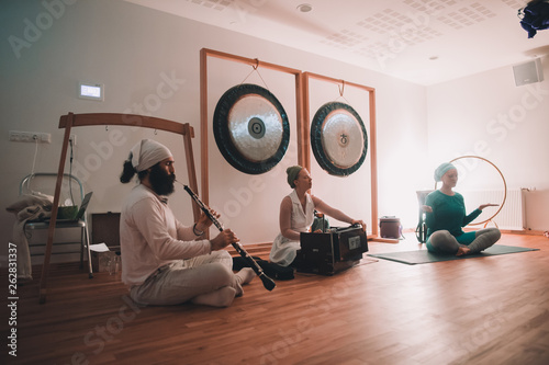Woman sitting in lotus pose near musicians playing on ethnic instruments near gongs in room