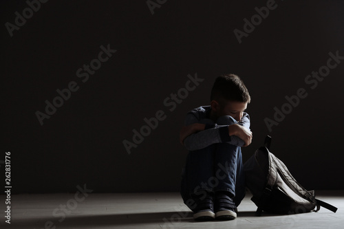 Upset boy with backpack sitting in dark room. Space for text