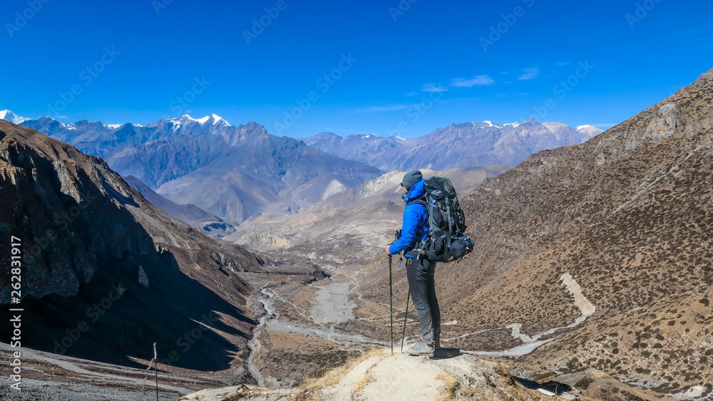 Man admires Himalayas, carries big backpack, supports himself with sticks, Annapurna Circuit Trek, Nepal. Upper Shreekharka. Dry grass. Socks dry on the backpack. Clouds in the mountains.