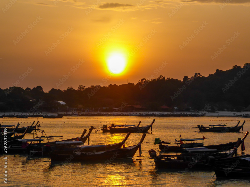 Dozens of boats anchored by the shore of the beach. The sun is going down behind the hill in the back. Soft orange colors of a sunset. Beach looks empty.
