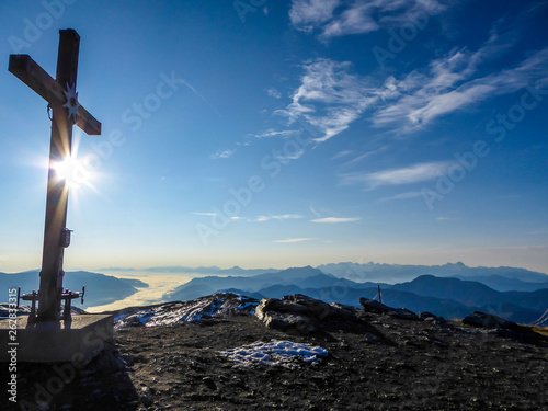 A cross placed at the top of a peak in Carnic Alps, Austria. Barren landscape with lots of rocks and pebbles on the grounds. Endless chains of mountains, covered with a bit of a mist.
