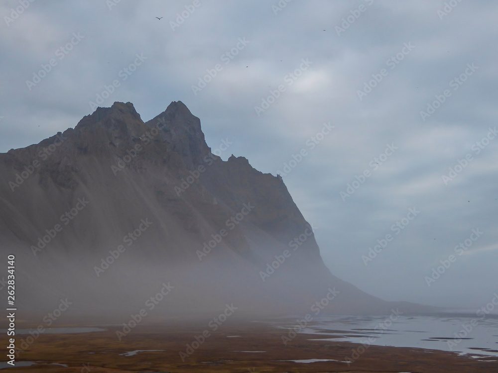 A chain of mountains emerging from the sea. Cloudy day. Shore is overgrown with golden grass. Some stones and pebbles on the shore as well. Mountains surrounded by mist, look splendid and glamorous.