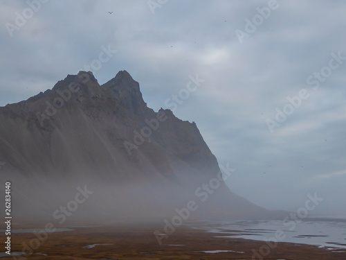 A chain of mountains emerging from the sea. Cloudy day. Shore is overgrown with golden grass. Some stones and pebbles on the shore as well. Mountains surrounded by mist, look splendid and glamorous.