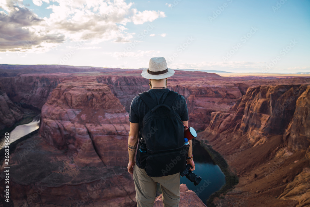 Back view of bearded guy with backpack holding photo camera to take pictures of beautiful canyon and calm river on sunny day on West Coast of USA Back view of bearded guy with backpack holding photo camera to take pictures of beautiful canyon and calm river on sunny day on West Coast of USA