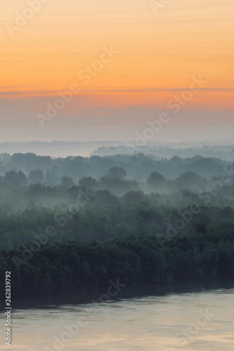 Mystical view on riverbank of large island with forest under haze at early morning. Mist among layers from tree silhouettes under predawn sky. Morning calm atmospheric landscape of majestic nature.